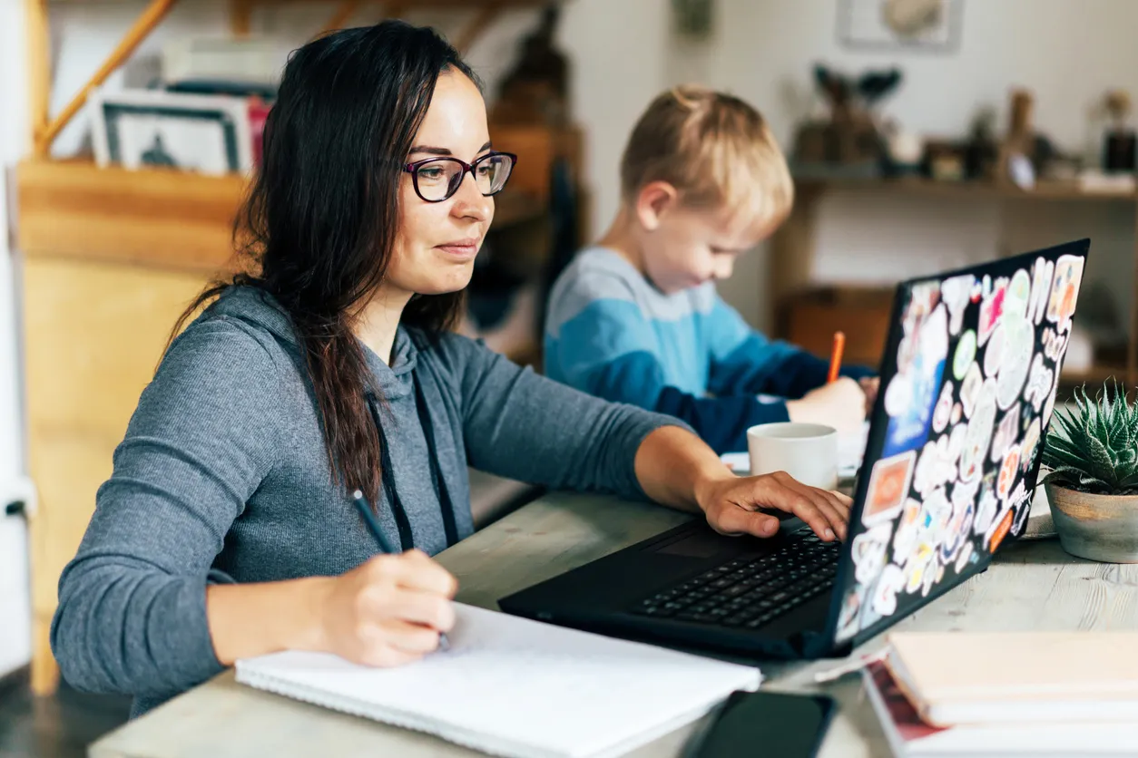 A parent reviewing documents on a laptop at home while their child sits alongside.
